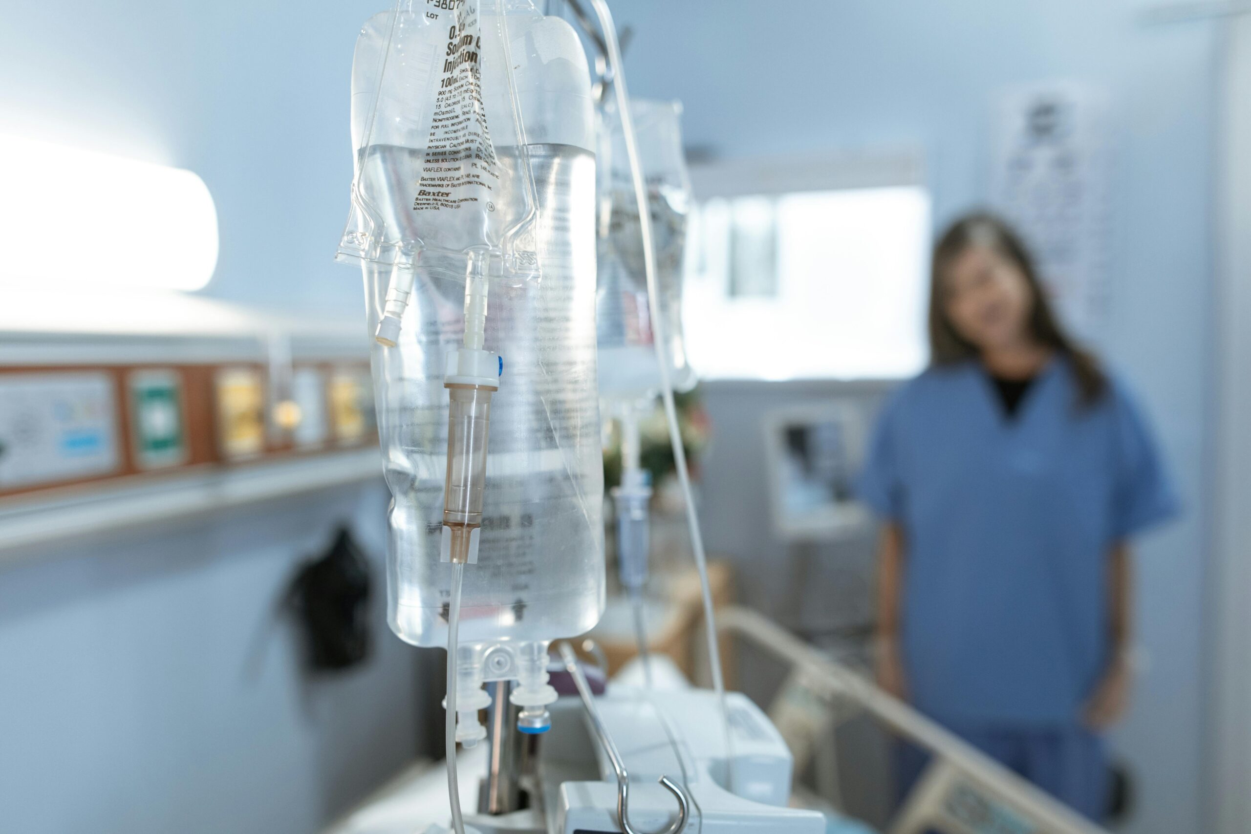 Close-up of an IV drip in a hospital with a blurred nurse in the background.